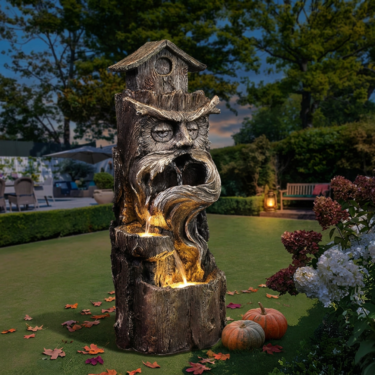 Illuminated tree-face water fountain with a birdhouse top beside pumpkins and autumn leaves at dusk